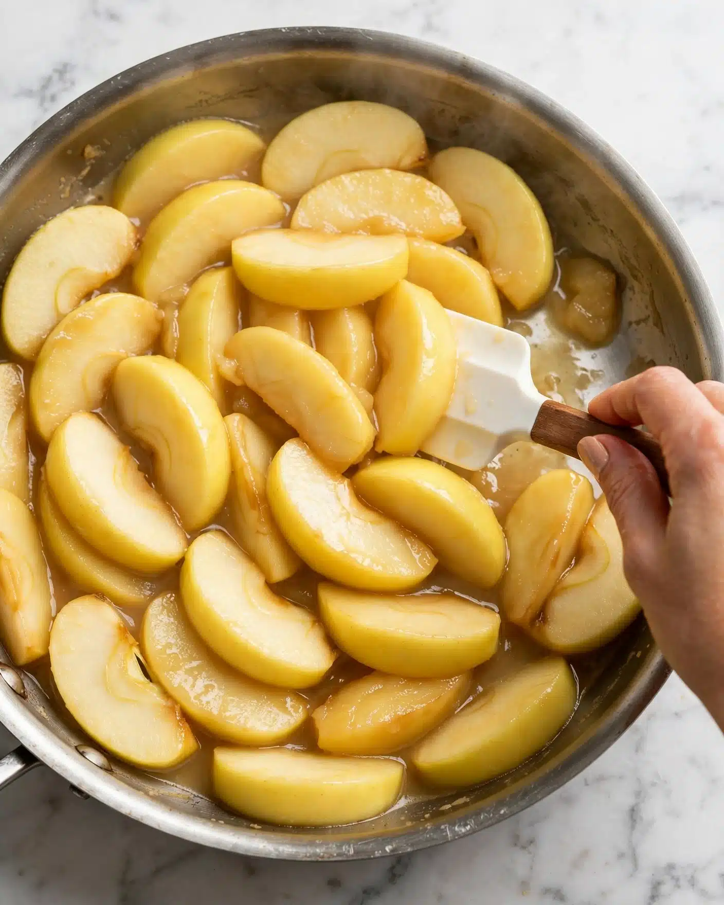 The image shows a close-up of a pan filled with many yellow apple slices being cooked. The apple slices are thick and soft-looking, covering the entire pan in a single layer. A white spatula with a brown handle is stirring the apples, held by a woman's hand at the right edge of the pan. The pan itself has a metallic shine, and the background is a white marbled surface. photo taken with an iphone --ar 4:5 --v 7