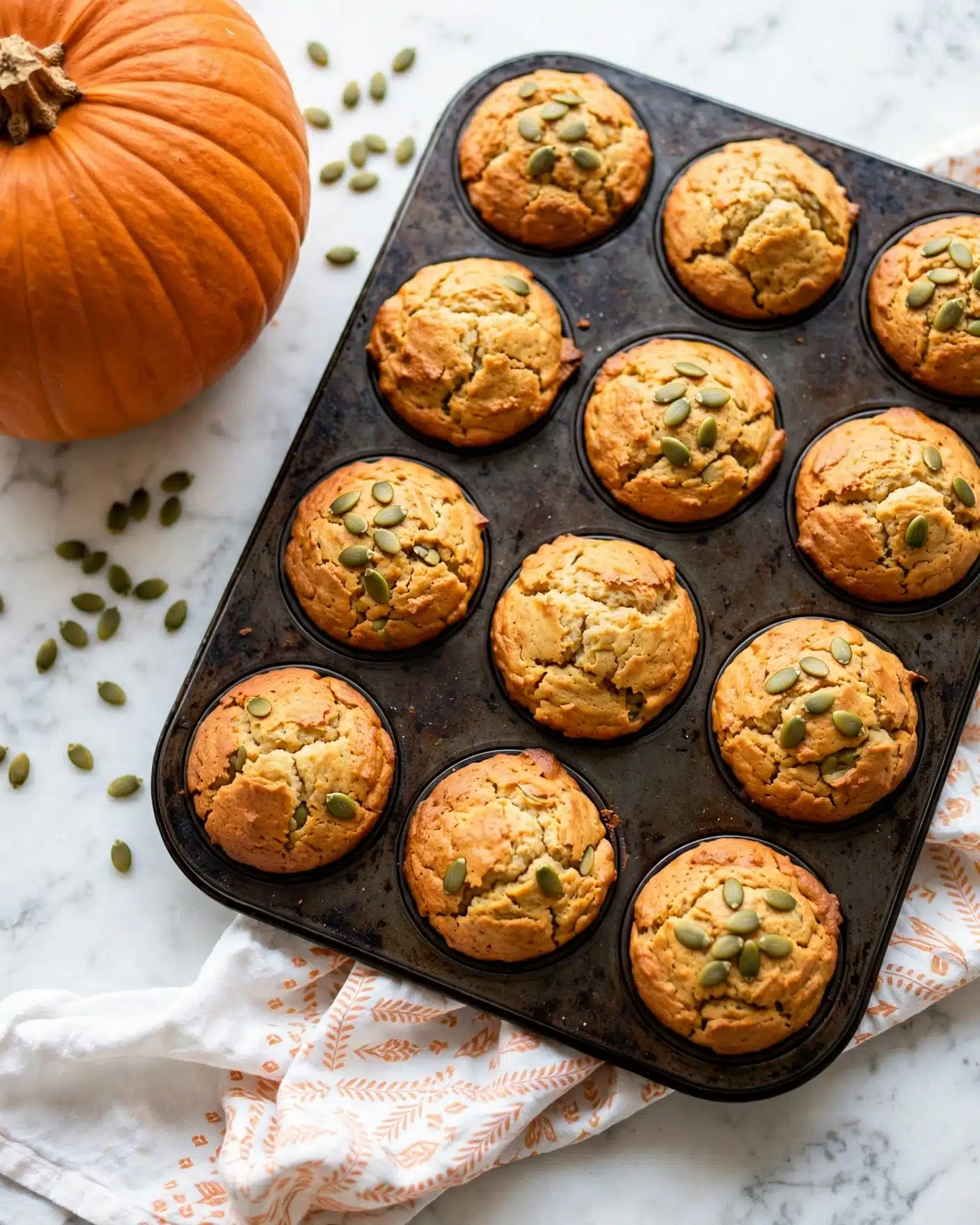Dairy-Free Pumpkin Muffins Recipe 4 The image shows a dark metal muffin pan filled with twelve golden-brown muffins, some topped with green pumpkin seeds arranged neatly on top. The muffins have a slightly cracked texture on the surface, giving a fresh-baked look. The pan rests on a white marbled surface with a patterned white and light orange cloth partially beneath it. To the left side, a large orange pumpkin and scattered pumpkin seeds are visible on the white marbled surface. The photo has bright, natural lighting emphasizing the warm colors of the muffins and pumpkin seeds. photo taken with an iphone --ar 4:5 --v 7