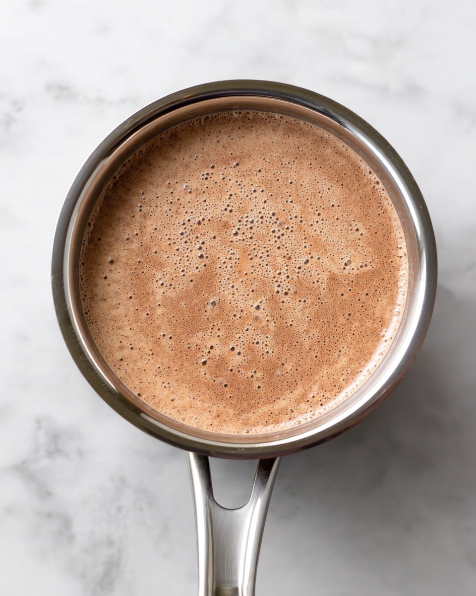 A top-down view of a shiny silver pot filled with a light brown liquid that has a smooth, foamy surface with tiny bubbles, showing a slightly speckled texture. The pot has a handle extending from the bottom middle and is placed on a white marbled textured surface. The liquid appears gently swirling, indicating it might be warming or mixing. photo taken with an iphone --ar 4:5 --v 7
