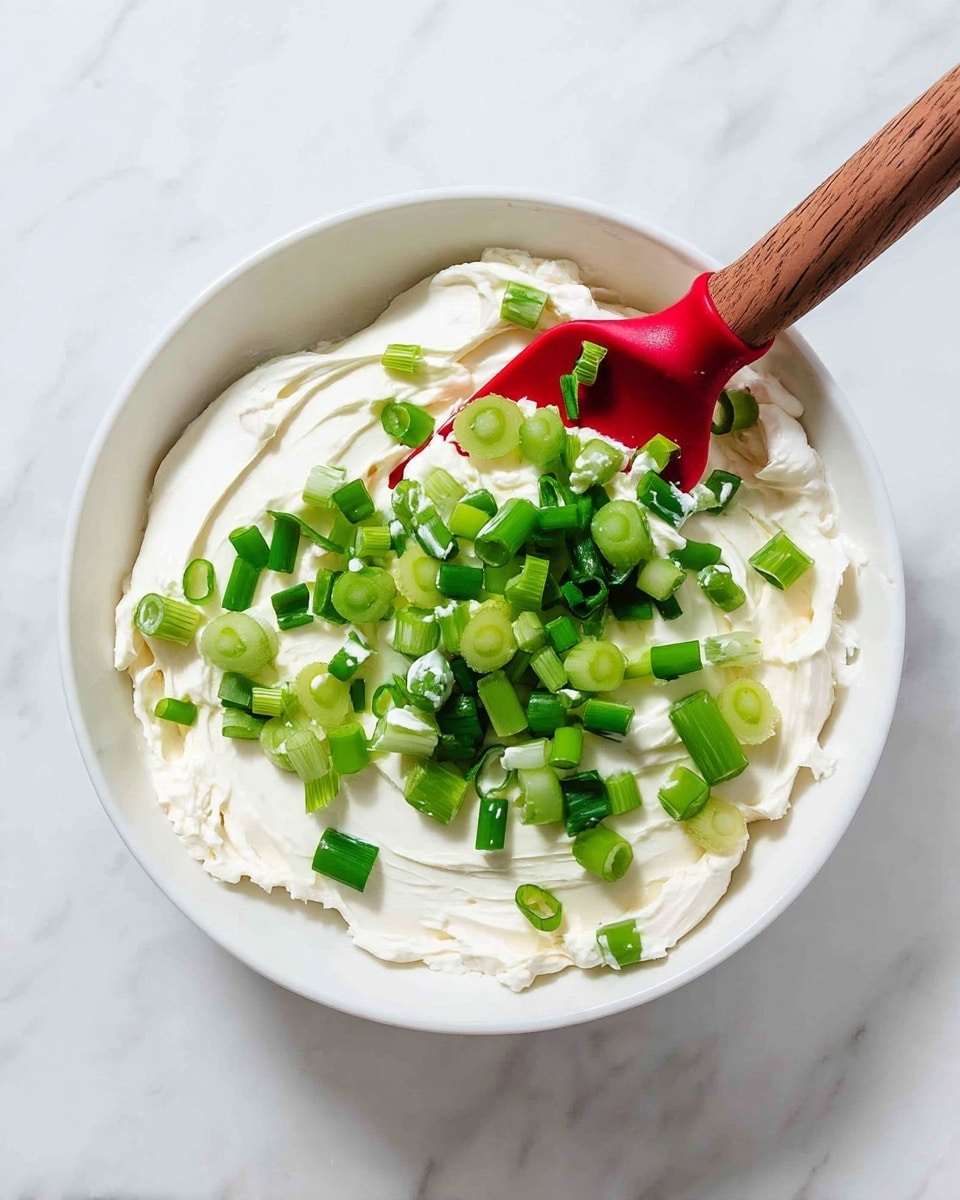 A white bowl is filled with a thick layer of smooth, creamy white spread, topped with a scattering of bright green chopped spring onions. A red spatula with a wooden handle rests on the spread, partially covered with the white creamy layer and green onions. The bowl is set against a white marbled background. photo taken with an iphone --ar 4:5 --v 7