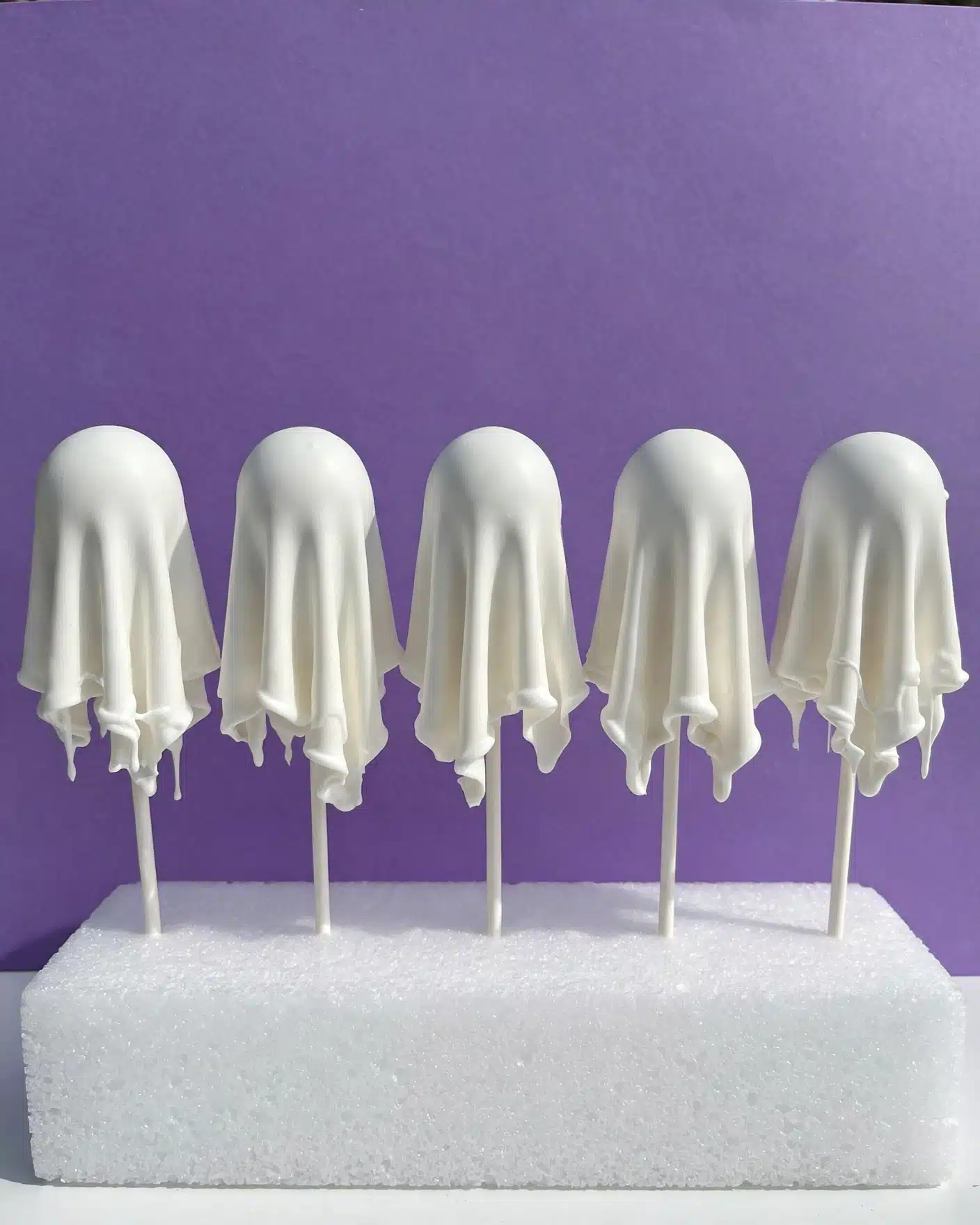 The image shows five ghost-shaped cake pops standing on white sticks, each covered in smooth and glossy white icing that drapes softly, creating a flowing ghost-like shape. They are positioned on a white foam block, with a plain purple background behind them, emphasizing their bright white color and smooth texture. The shapes have small dips and ripples at the bottom edges, giving a natural fabric look. The overall scene is simple, with the cake pops evenly spaced and gently lit, highlighting their smooth surfaces and clean white color. photo taken with an iphone --ar 4:5 --v 7