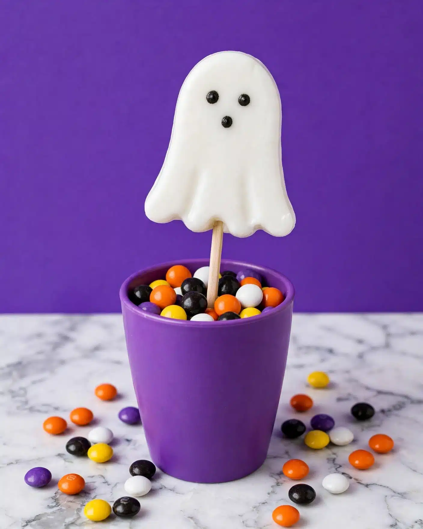 A white ghost-shaped candy pop on a stick stands upright in a vibrant purple cup filled with small, colorful round candies in colors like orange, black, yellow, white, and purple. The ghost has three black dots for eyes and mouth. The cup sits on a white marbled surface scattered with the same multicolored candies around it, with a solid purple background behind the cup. photo taken with an iphone --ar 4:5 --v 7
