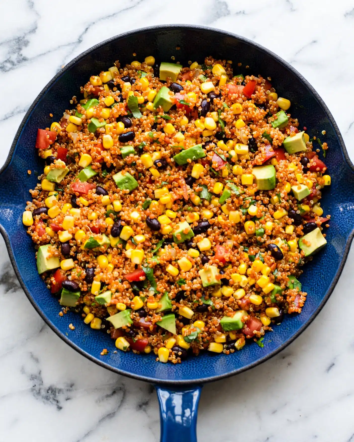 One Pan Mexican Quinoa Recipe 5 A blue cast iron pan filled with a colorful quinoa salad on a white marbled surface. The salad has small round grains of orange quinoa mixed with bright yellow corn kernels, black beans, green avocado cubes, and bits of red tomato. The ingredients are evenly spread and mixed, showing a fresh and textured look. The pan has a small blue handle visible in front. photo taken with an iphone --ar 4:5 --v 7