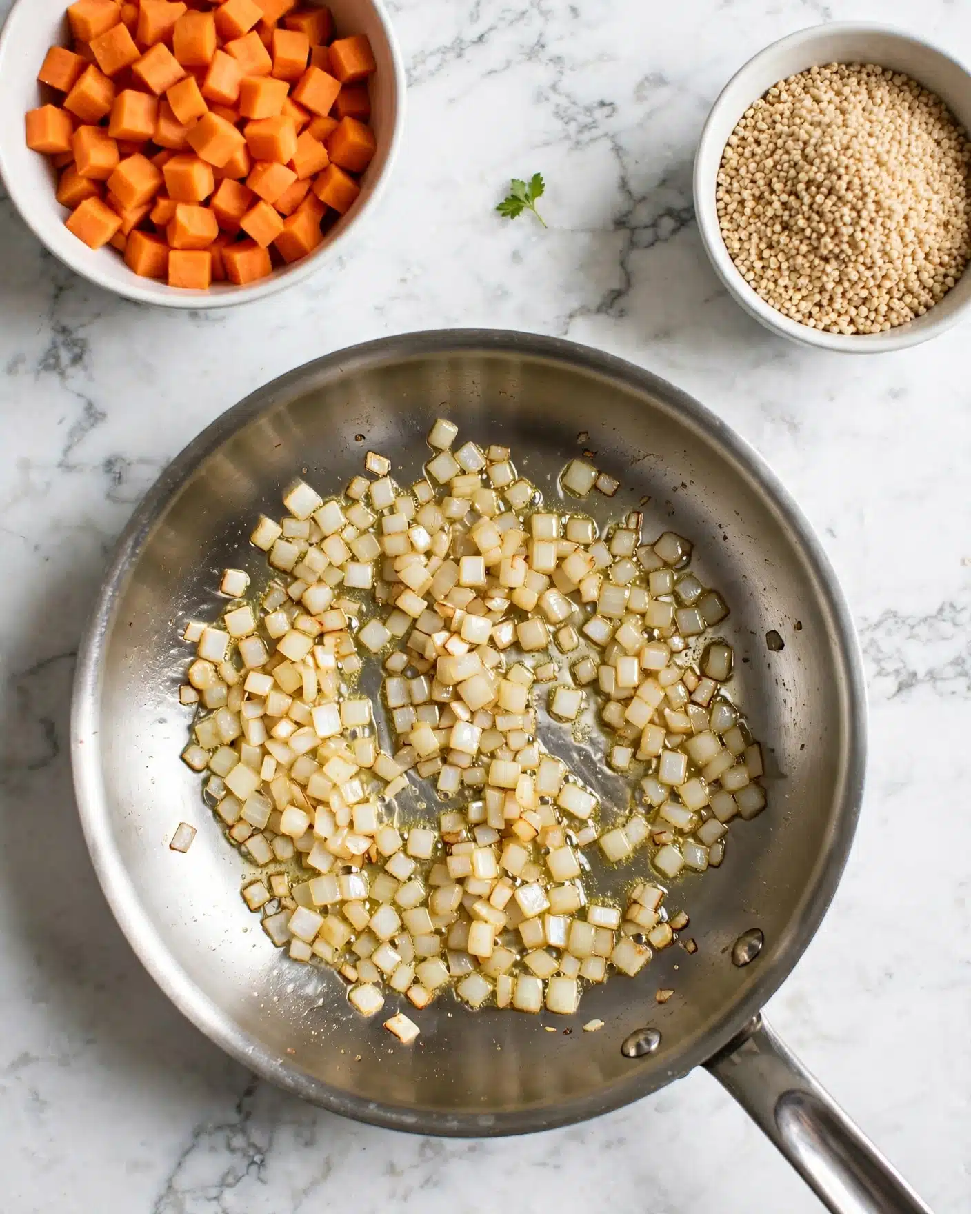 One Pan Mexican Quinoa Recipe 4 The image shows a silver frying pan on a white marbled surface, filled with small, light golden diced pieces that look like onions, cooking in oil. At the top left, there is a white bowl filled with small, orange cubed pieces, possibly carrots or sweet potatoes. At the top right, there is another white bowl filled with small, light brown grains, possibly quinoa or couscous. A small green herb leaf lies on the surface between the two bowls, adding a touch of color. Photo taken with an iphone --ar 4:5 --v 7