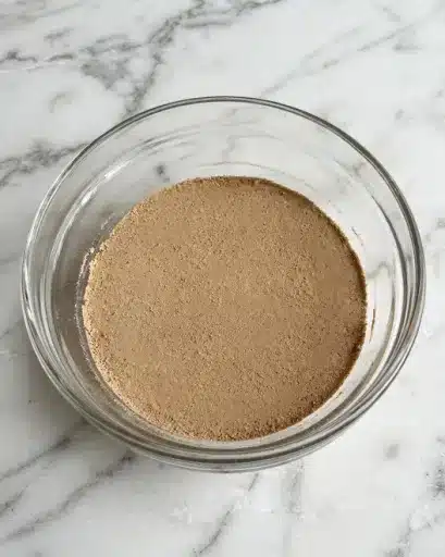 A clear glass bowl sits on a white marbled surface, filled with a single layer of fine, light brown powder with a slightly gritty texture. The powder spreads evenly, covering the bottom of the bowl in a thin, smooth layer without any visible lumps or separation. In the top left corner, a white circle with the black number 